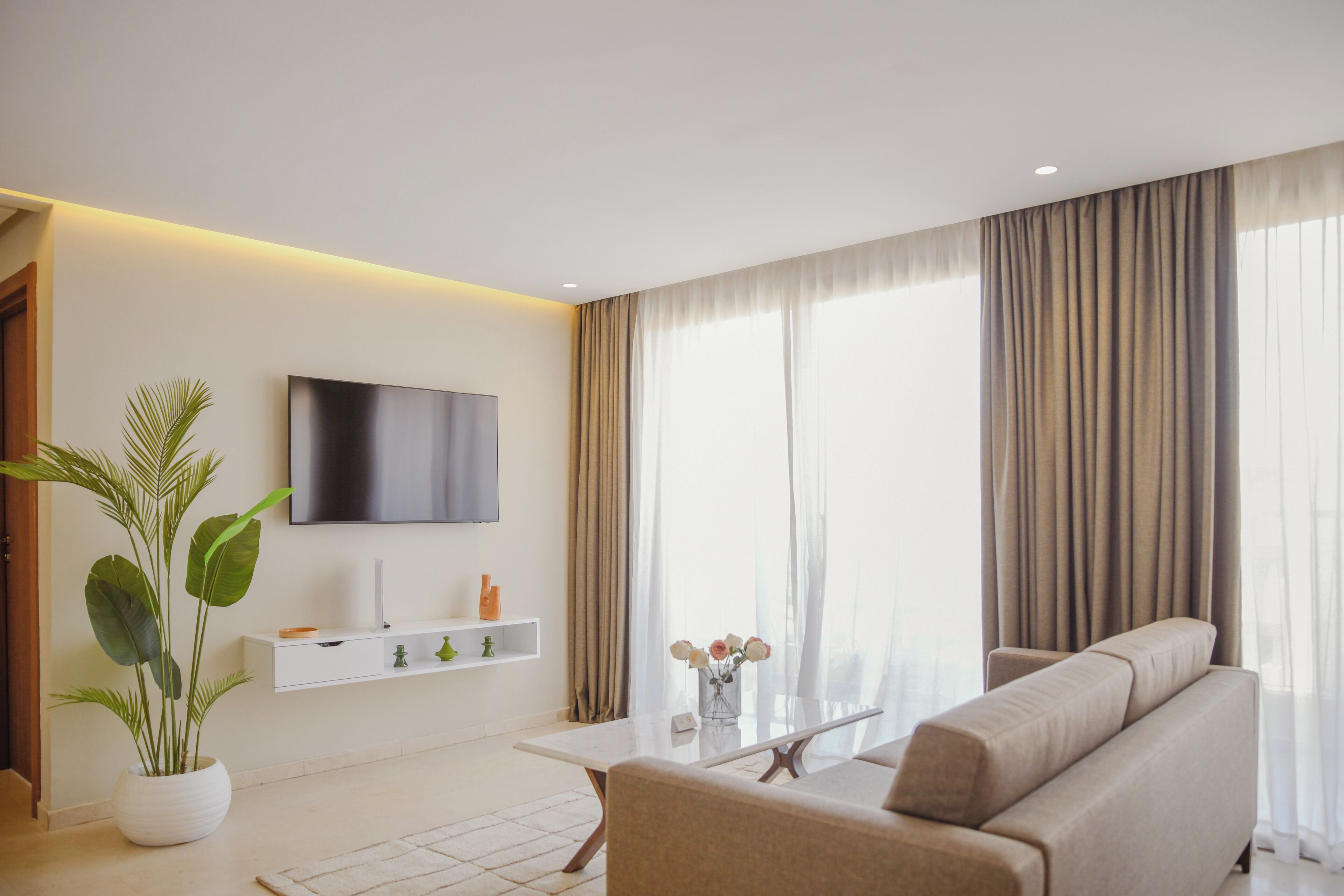 Una sala moderna con televisor de pantalla plana, un sofá, una mesa de centro, una planta en maceta y una ventana con cortinas.
