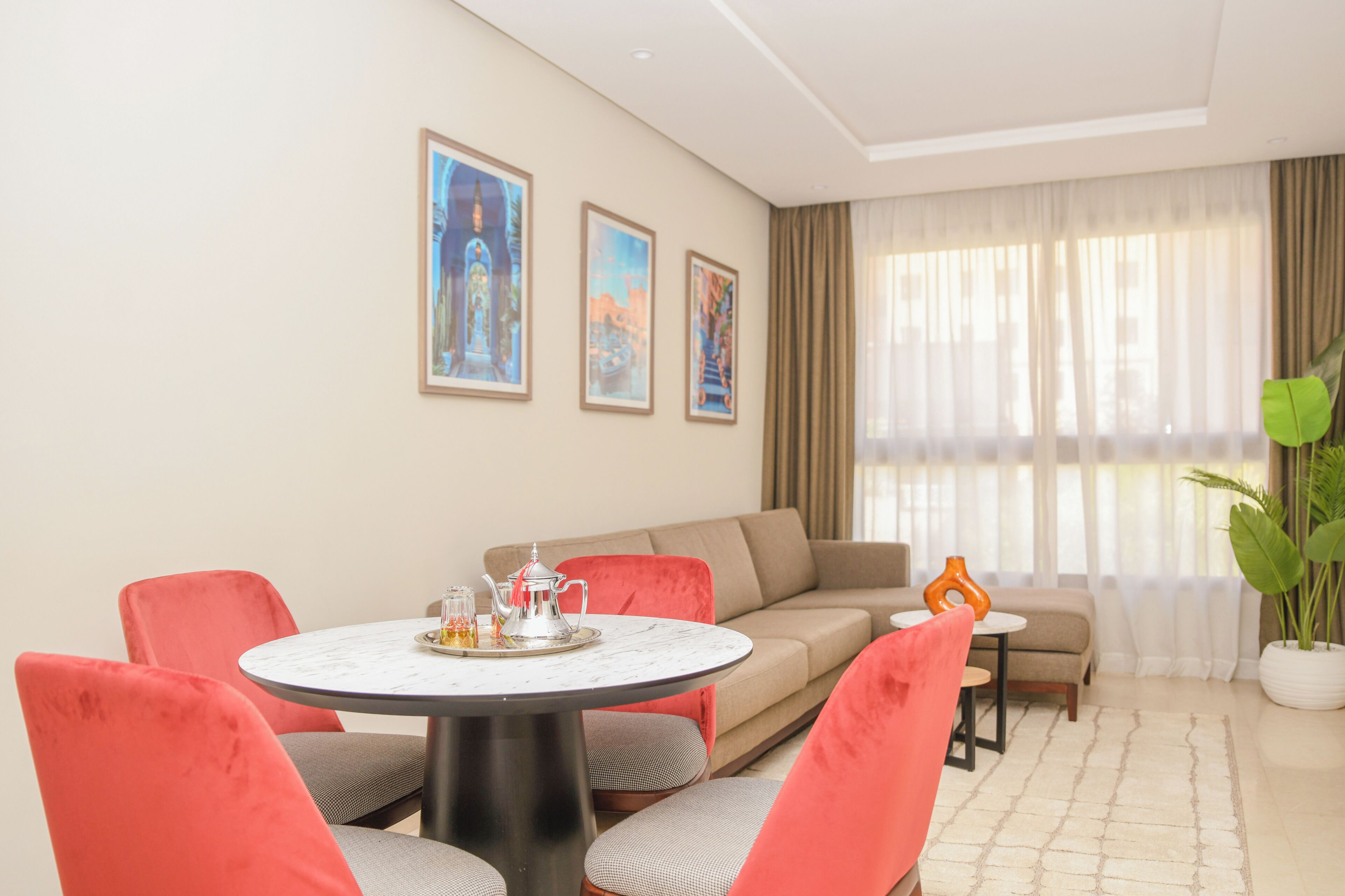A modern living room with a round dining table, red upholstered chairs, a beige sofa, and two framed artworks on the wall.
