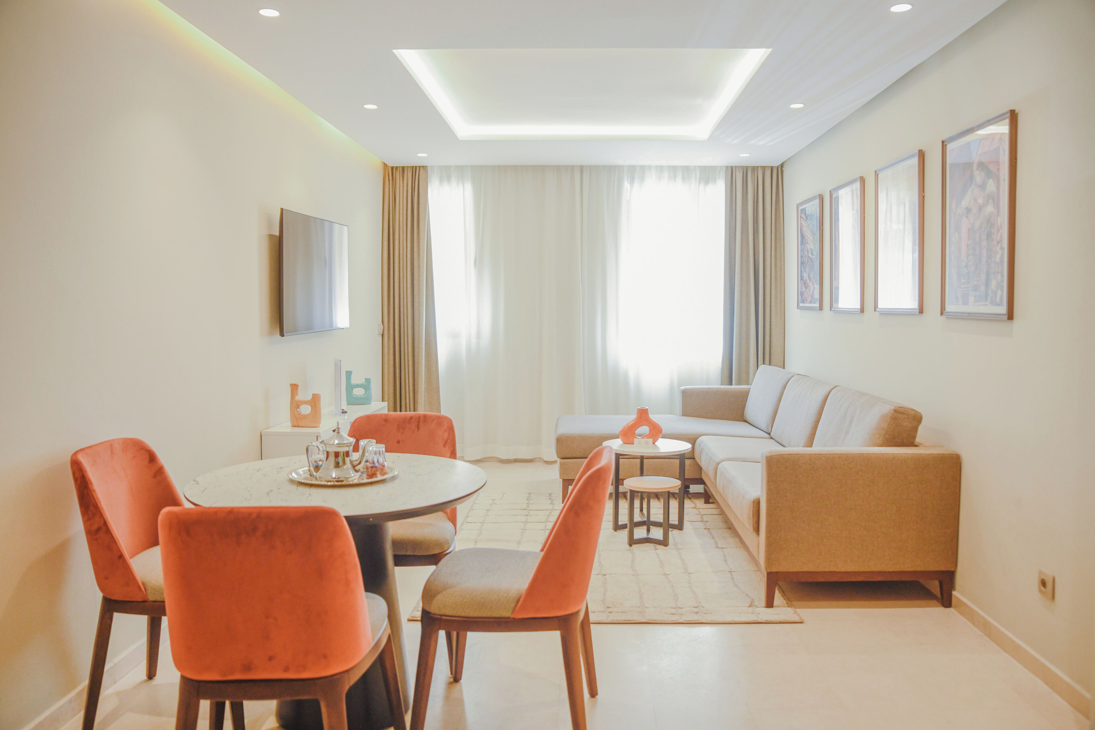 A modern living room with a dining area, featuring a sofa, chairs, a coffee table, and a flat-screen TV mounted on the wall.