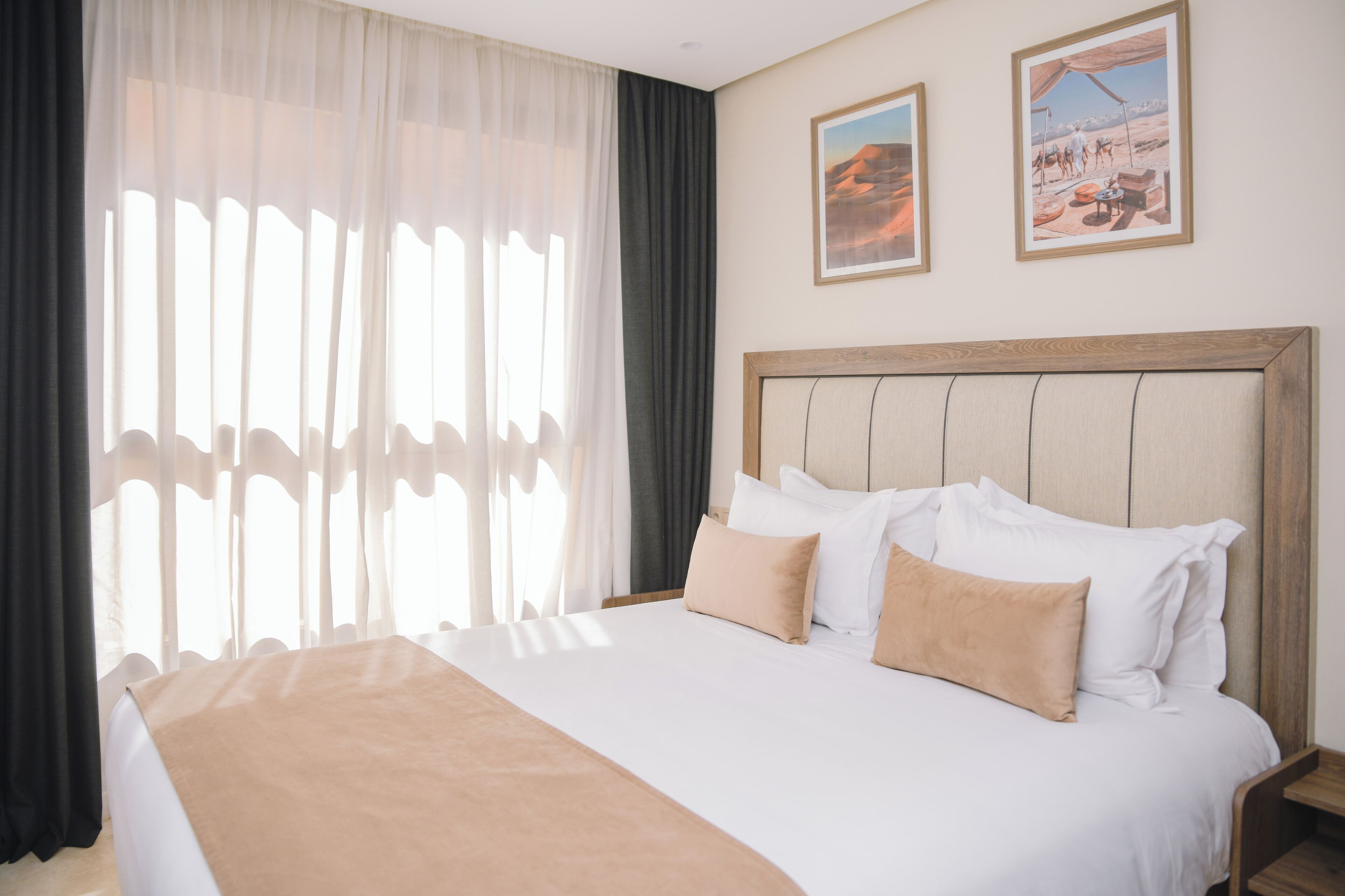 A hotel room with a large bed, two paintings on the wall, and sheer curtains.