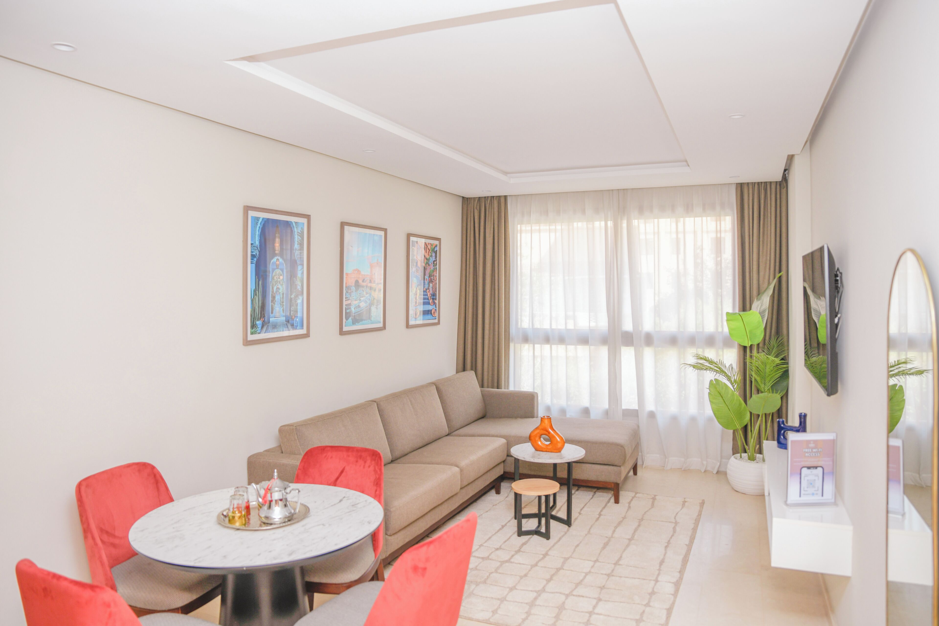 Una sala de estar moderna con un sofá beige, una mesa de centro redonda y sillas rojas. Hay tres cuadros enmarcados en la pared, un televisor montado y una planta en maceta.