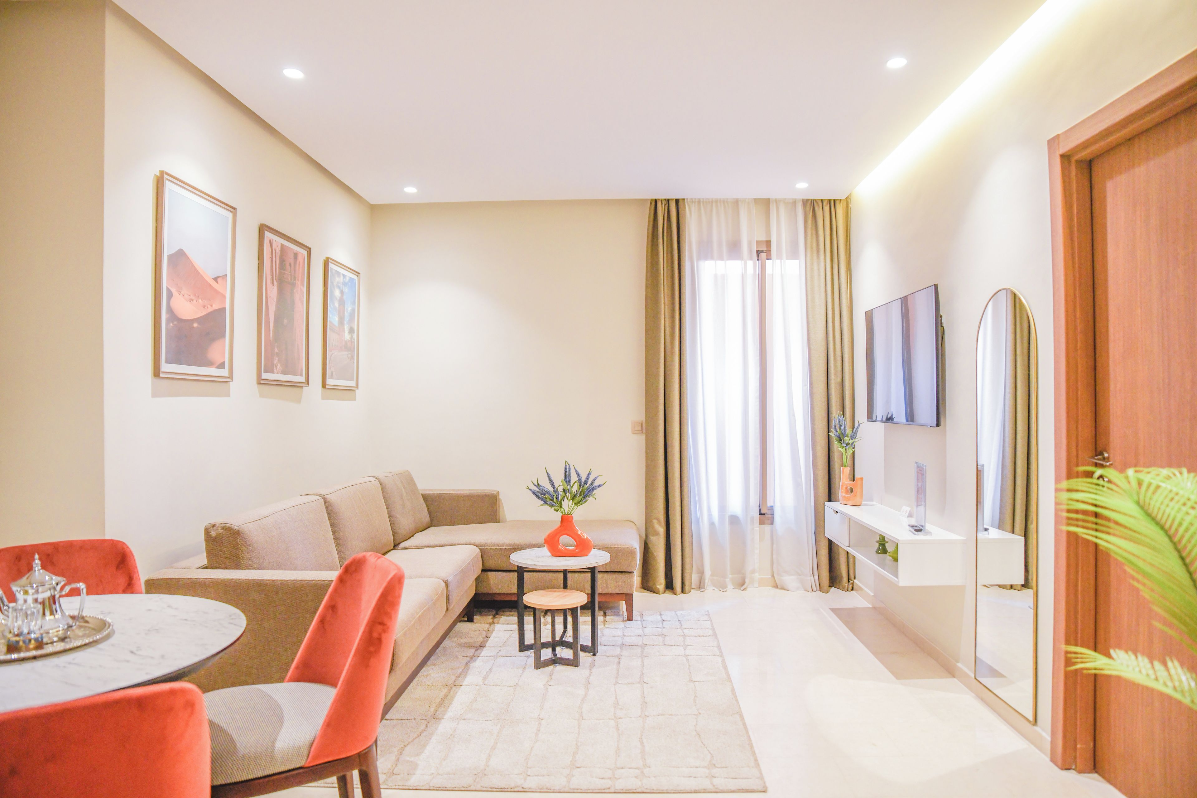 A modern living room with a sofa, a dining table with chairs, a TV mounted on the wall, and a small side table with a vase.