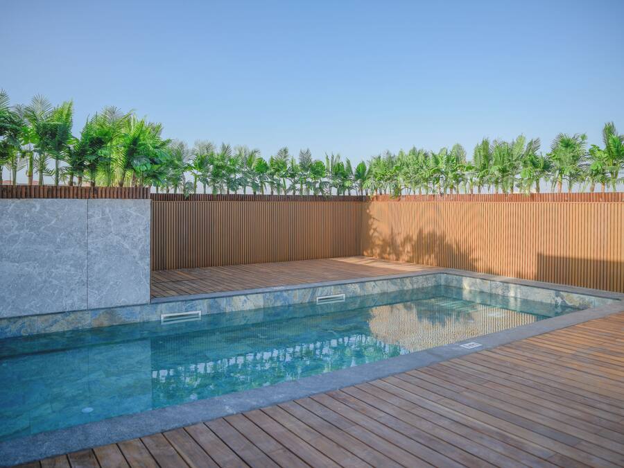Piscina con muelle de madera y pared de piedra, rodeada por una valla de madera y palmeras.
