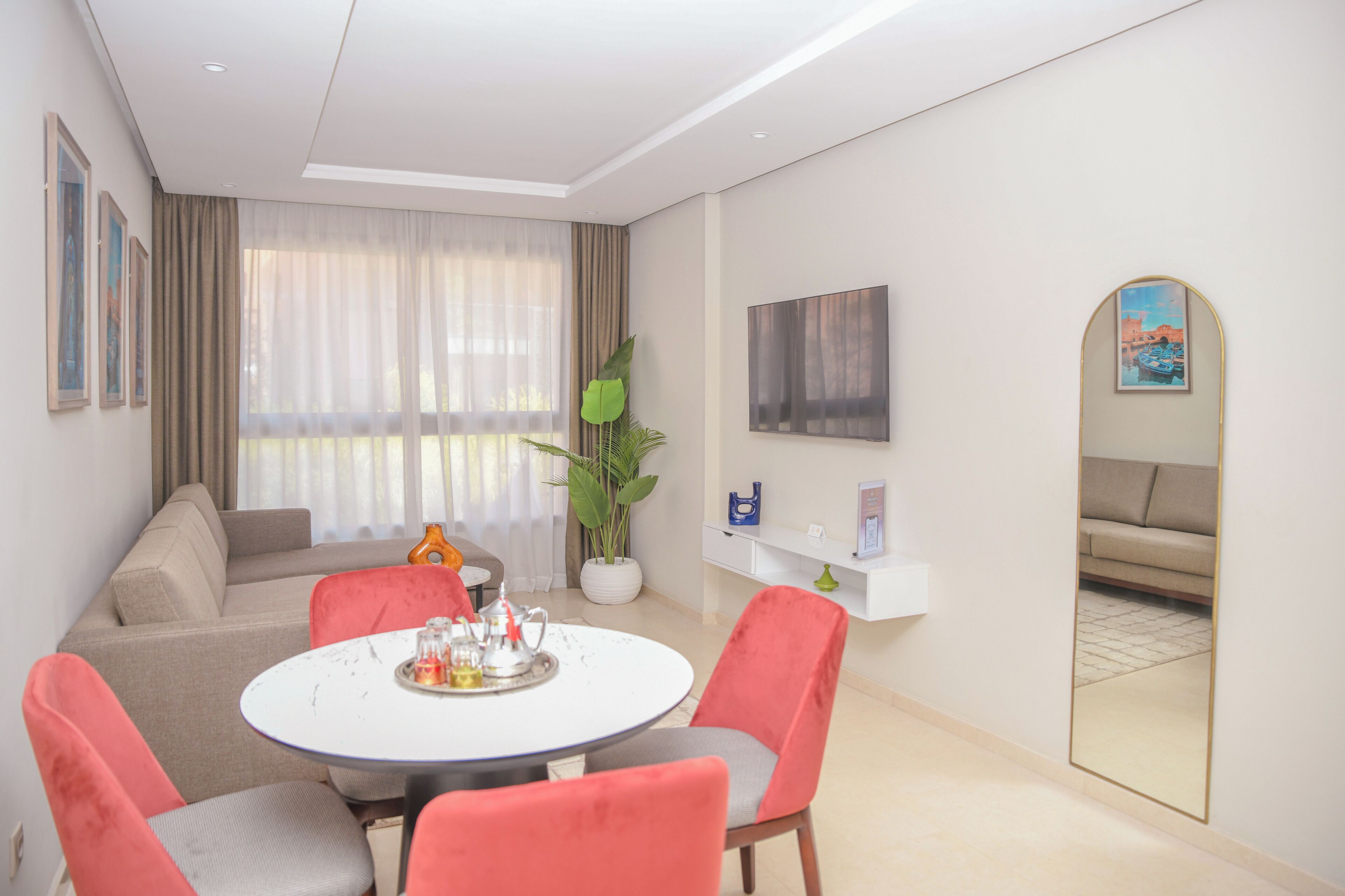 Ein modernes Wohnzimmer mit einem runden Esstisch, roten Stühlen, einer Couch, einem großen Fenster mit Vorhängen, einem Topf mit Pflanze, einem Fernseher und einem Spiegel.