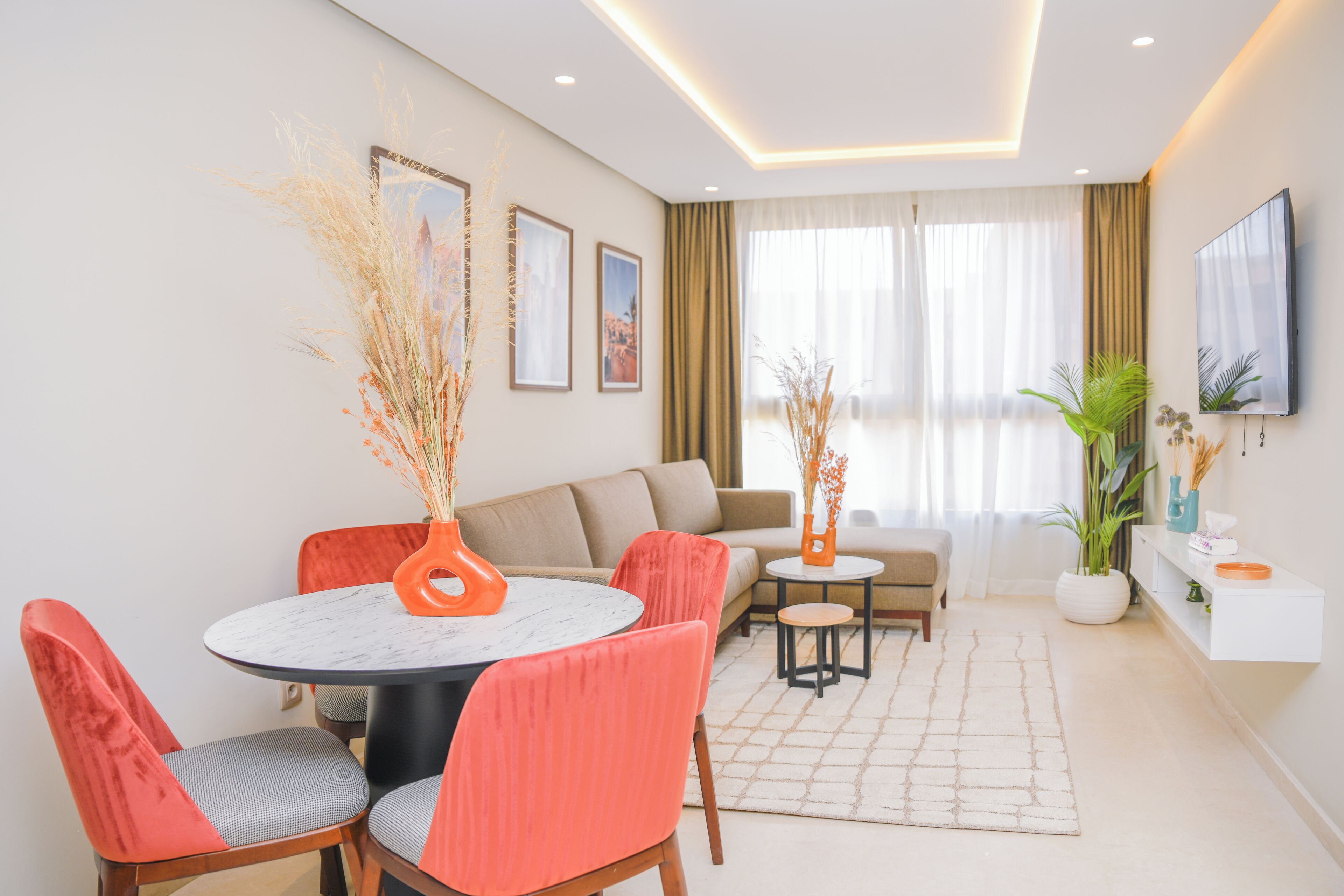 A modern living room with a round dining table, red chairs, a sofa, a TV mounted on the wall, and a potted plant.