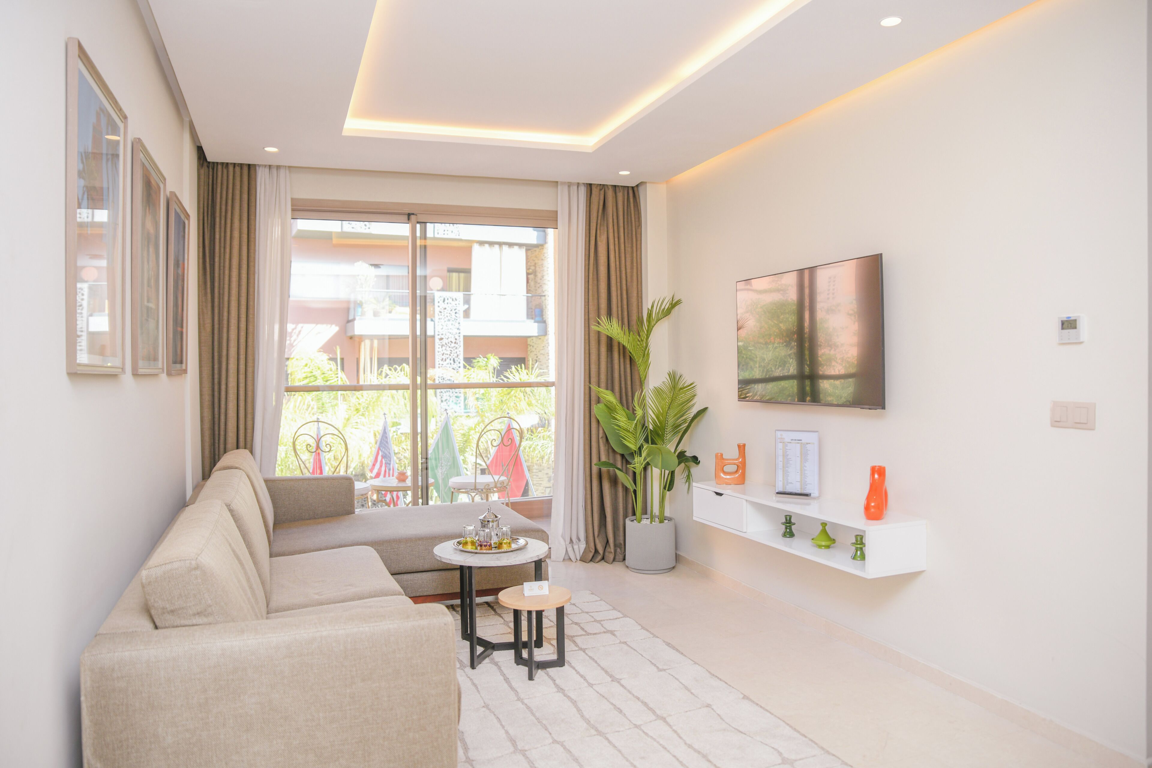 Un salon moderne comprenant un canapé, une petite table ronde, une plante en pot, un téléviseur à écran plat fixé au mur et un balcon aménagé avec du mobilier d’extérieur.