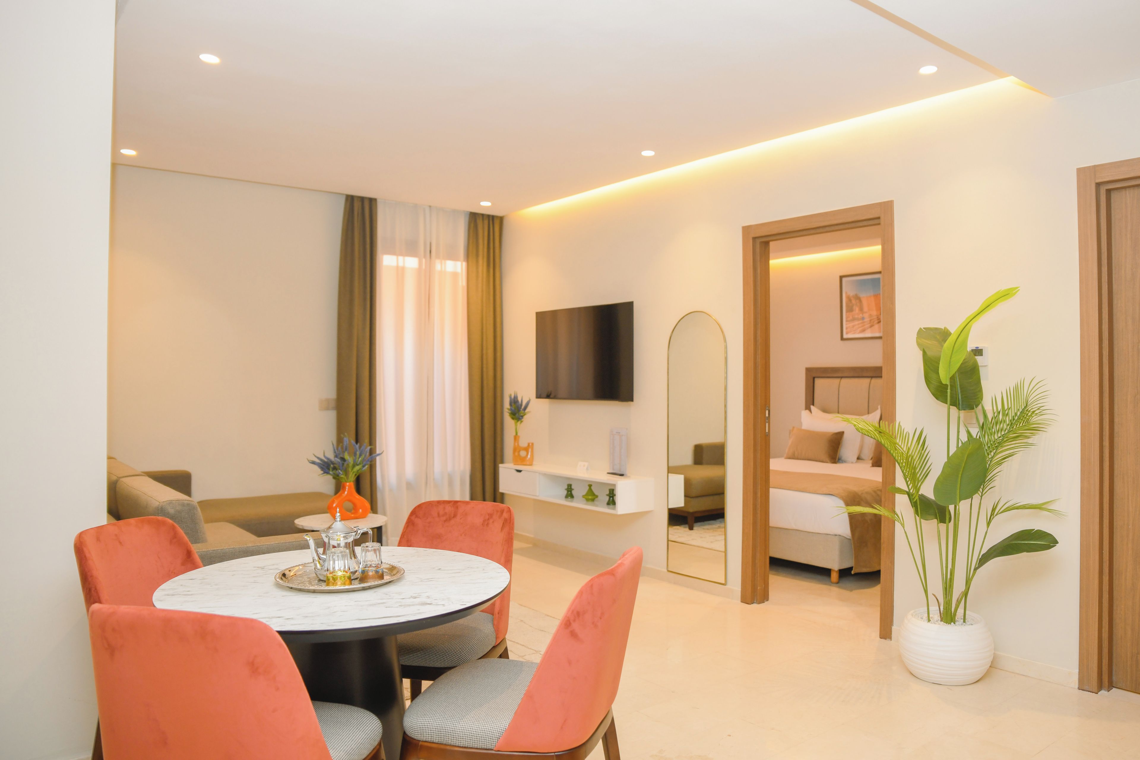 A modern living room with a round dining table, orange chairs, a sofa, a TV mounted on the wall, and a large mirror.