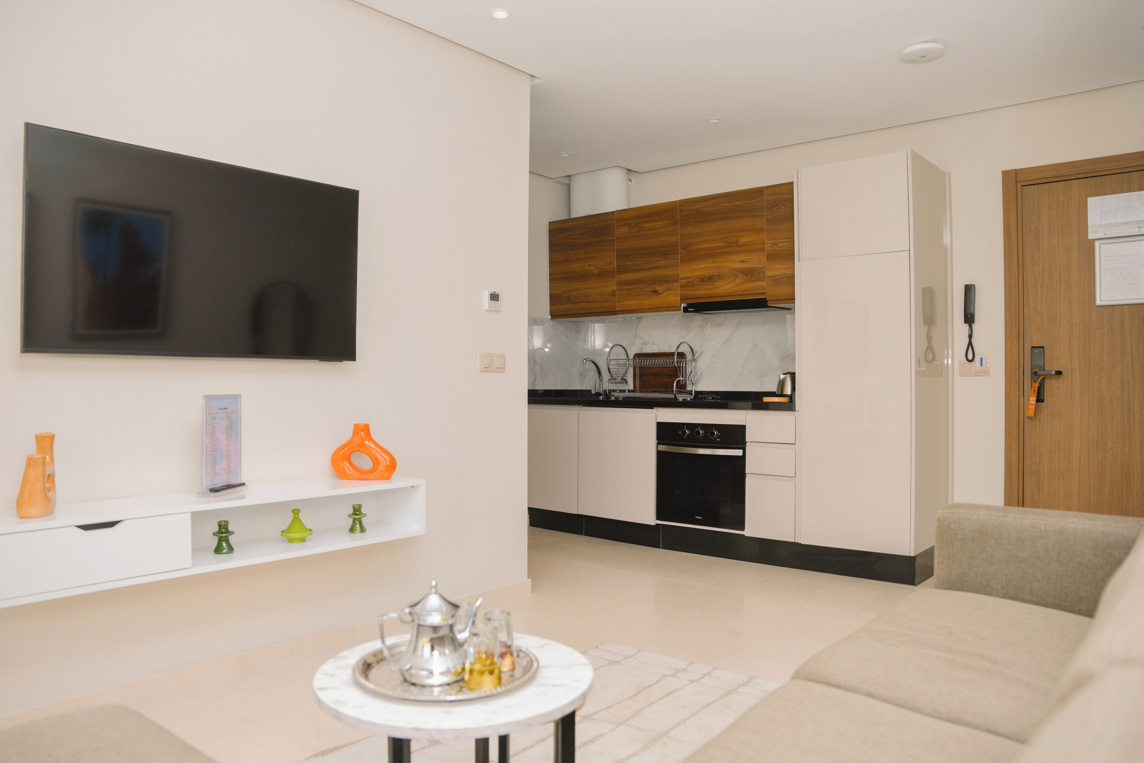 A modern living room with a flat-screen TV, a coffee table with decorative items, a sofa, and a kitchen area with wooden cabinets and a built-in oven.