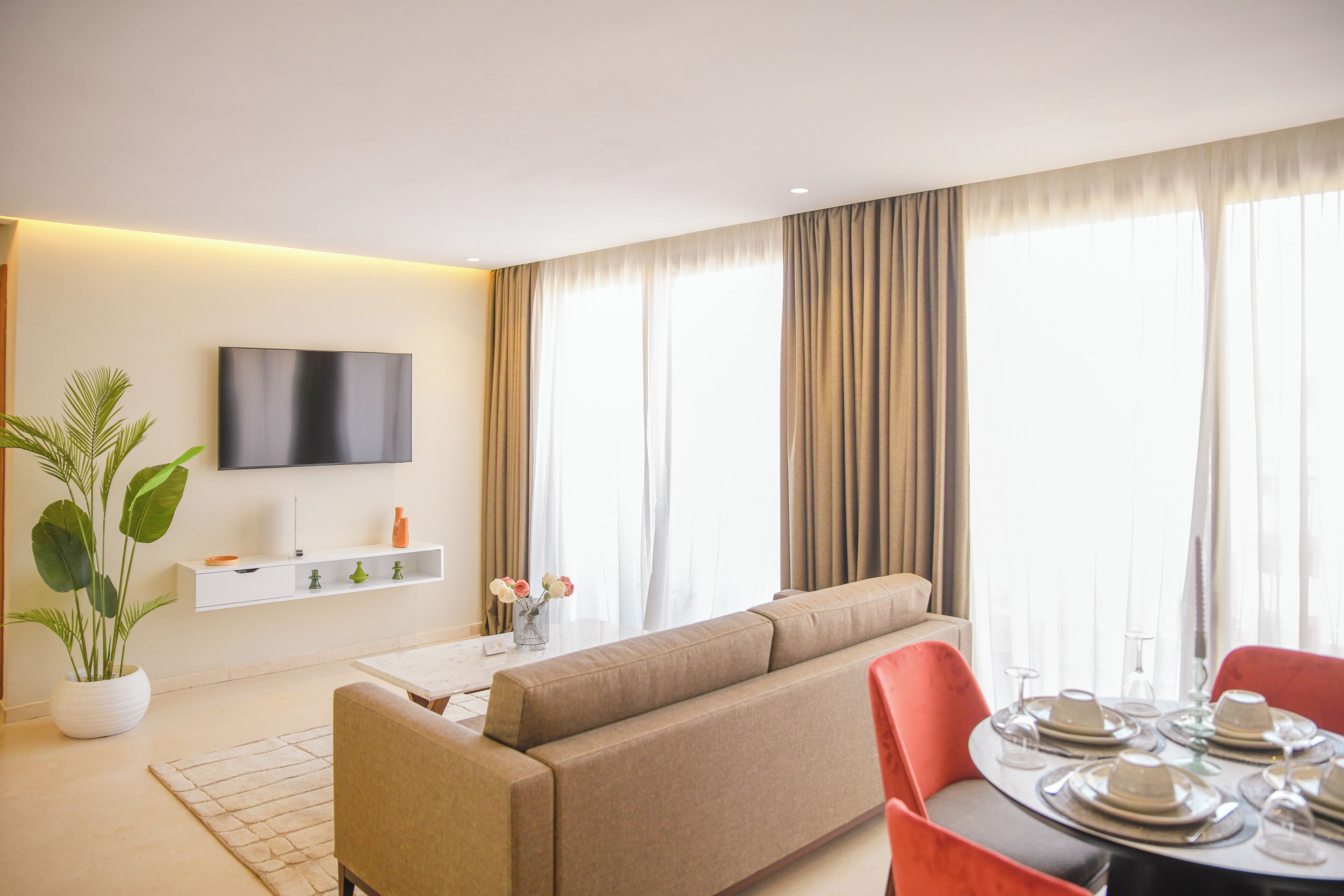 A modern living room with a sofa, a flat-screen TV, a dining table set for four, and a large window with curtains.