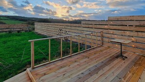 Um deck de madeira com guarda-corpo, com vista para um campo verde e um horizonte distante.