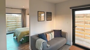 Uma sala de estar moderna com um sofá cinzento, uma cama com colcha verde e uma grande porta de vidro deslizante que dá para uma área externa.