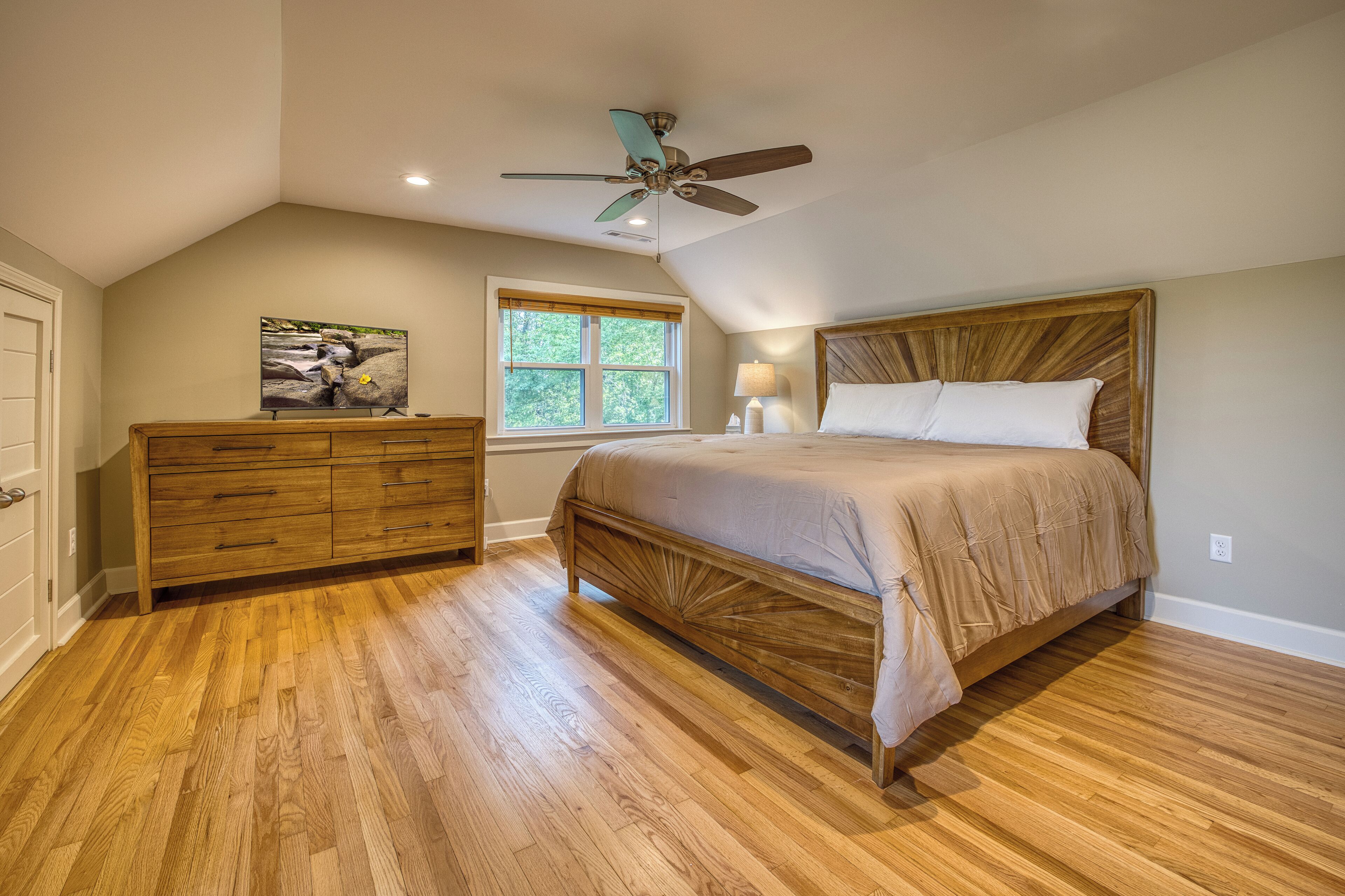 Quarto com cama de madeira, cômoda, ventoinha no teto e janela com vista para árvores.