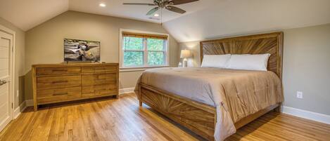 Quarto com cama de madeira, cômoda, ventoinha no teto e janela com vista para árvores.