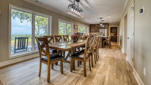 Un comedor amplio con una larga mesa de madera y sillas, ventanas grandes que ofrecen vistas al exterior, y una cocina moderna al fondo.
