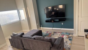 A living room with a grey sofa, a flat-screen TV mounted on the wall, a patterned rug, and a door to the right. - Modern 2BR/2BTH near DC; Walk to Mall, Shops & Metro! (Hyattsville)
