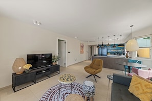 A modern living room with a TV, sofa, and coffee table. There is a kitchen area with bar stools and a dining table. - The Strand apartment 1A, 5 guests with pool and beach (Willemstad)