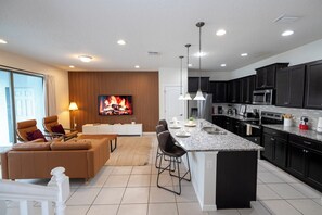 A modern kitchen with dark cabinets, a central island, and a flat-screen TV mounted on a wooden wall. - Ideal Family 5BR Villa in Windsor at Westside Resort ● Private Pool, BBQ & more! (Kissimmee)