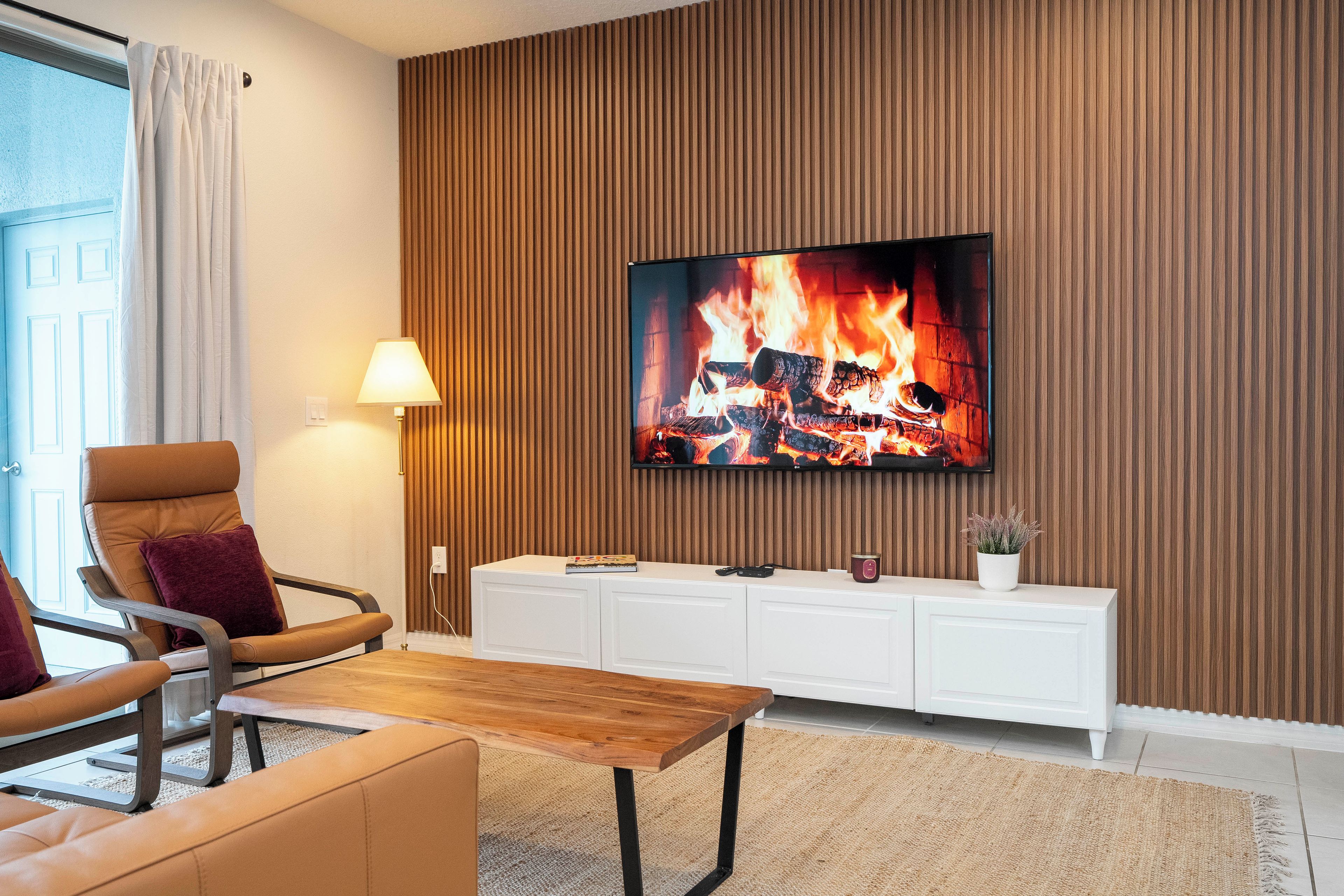 A modern living room with a flat-screen TV mounted on a wooden panel wall, a leather armchair, a wooden coffee table, and a white console unit with a plant and a candle.