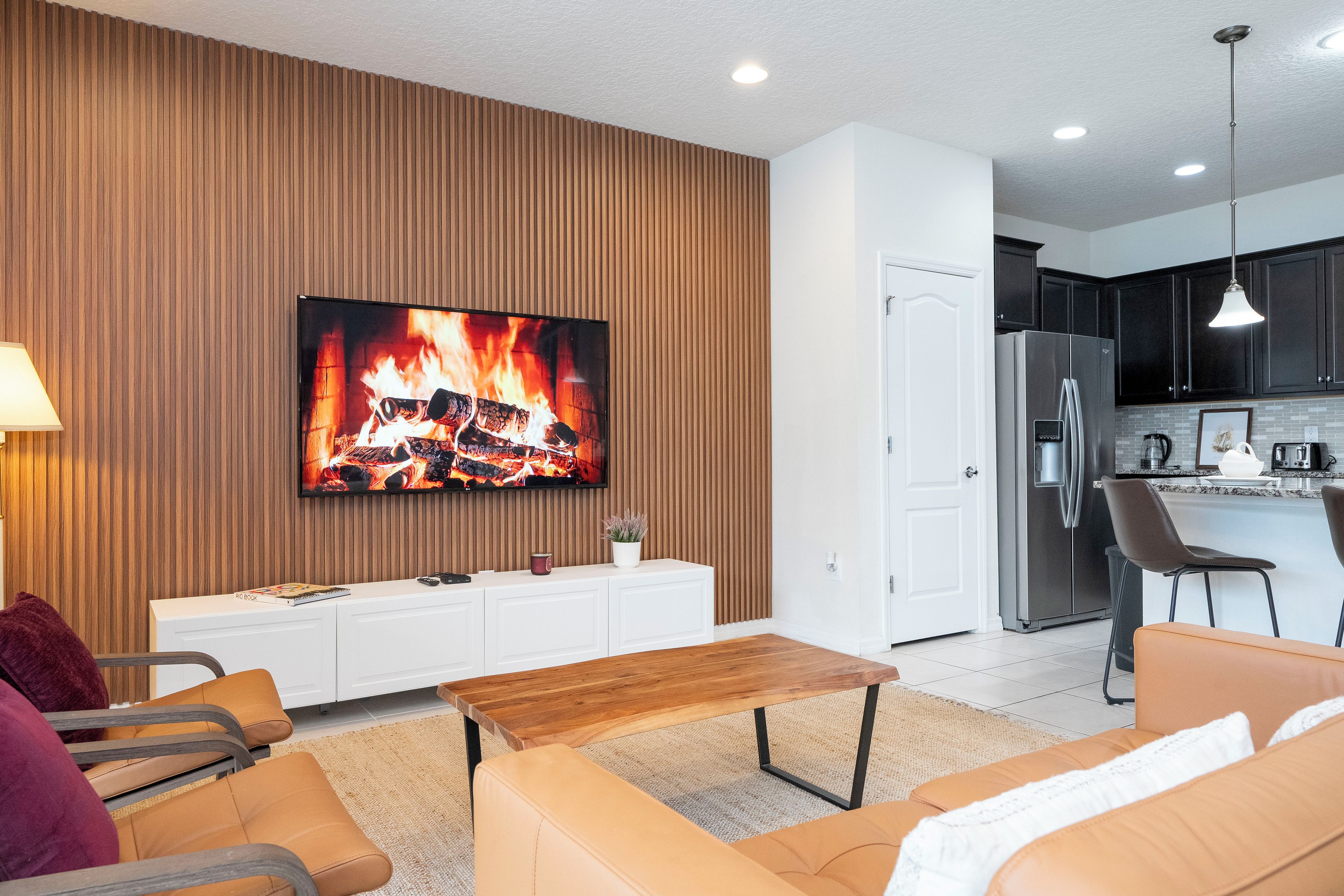 A modern living room with a flat-screen TV, a wooden accent wall, a sofa, a coffee table, and a kitchen area with a refrigerator and cabinets.