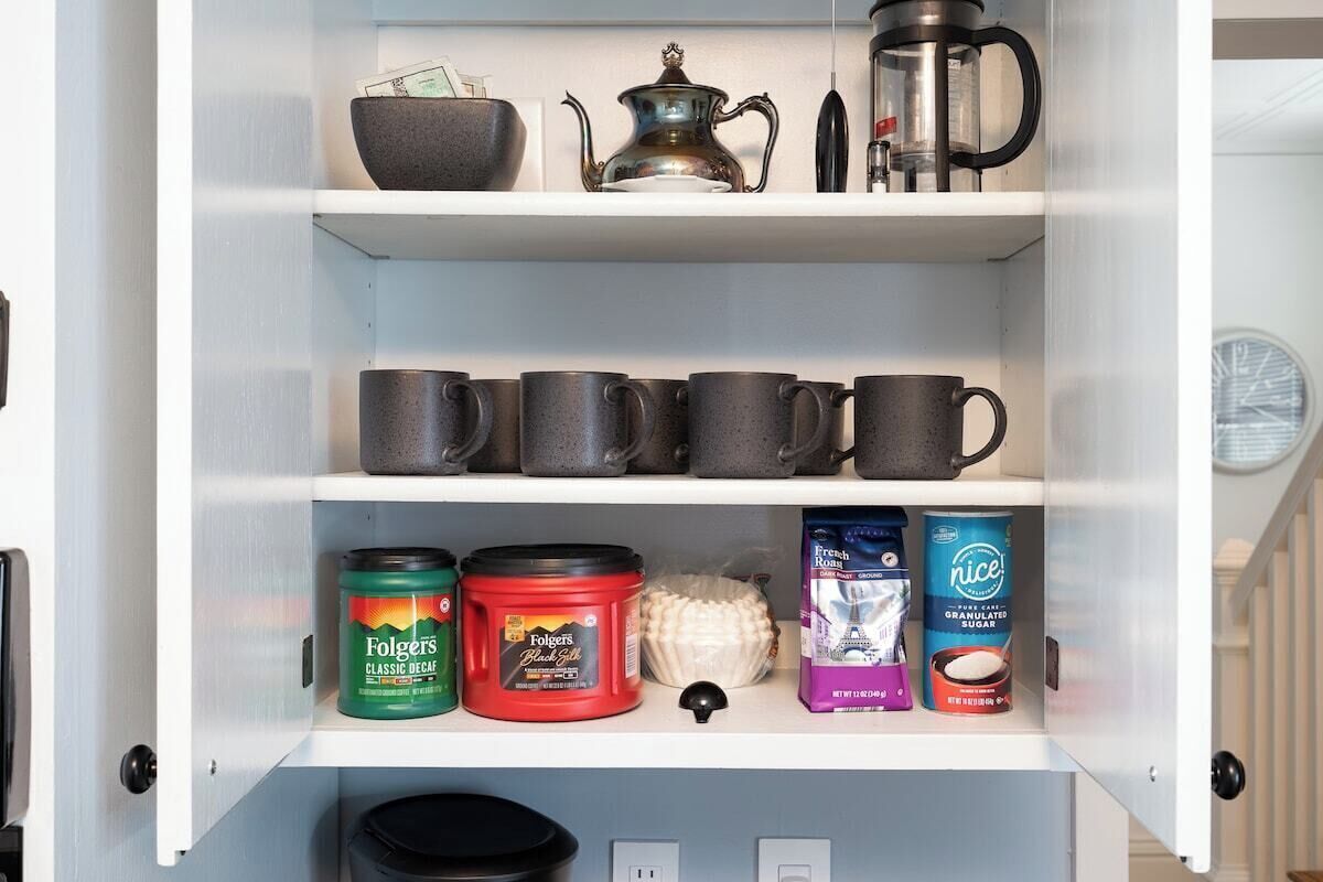A kitchen pantry with shelves containing mugs, Folgers coffee, Folgers black tea, French roast coffee, and Nice granulated sugar.