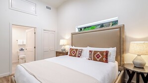 Ein Hotelzimmer mit einem großen Bett, einem Nachttisch mit Lampe, einem Badezimmer mit Toilette und Dusche sowie einem Blick ins Freie durch ein Fenster.