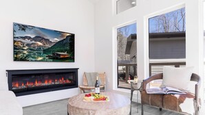 Ein modernes Wohnzimmer mit Kamin, einem runden Couchtisch mit Obst und Käse, einem bequemen Sessel und einem großen Landschaftsbild in einem Rahmen.