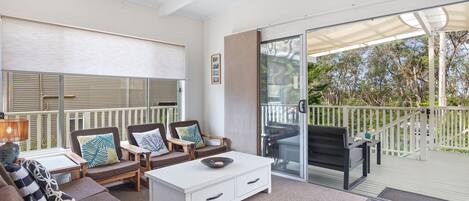 A modern outdoor living area with a white sofa, two armchairs, a coffee table, and a dining table. There is a sliding glass door leading to a balcony with a view of trees.