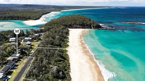 Eine Vogelperspektive einer Küstenlandschaft mit Strand, einer bewaldeten Halbinsel und einem Wohngebiet mit Häusern und einer Straße.