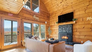 A cozy cabin interior with a fireplace, wooden walls, a sofa, and large windows offering a mountain view. - Best views in Cherry Log, Putting Green, Hot Tub (Cherry Log)