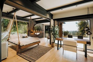 A modern bedroom with a hanging bed, a dining area with a wooden table, and large windows offering a view of the outdoors. - The Amalfi Glass Sanctuary – LA’ Riviera Coast (Los Angeles)