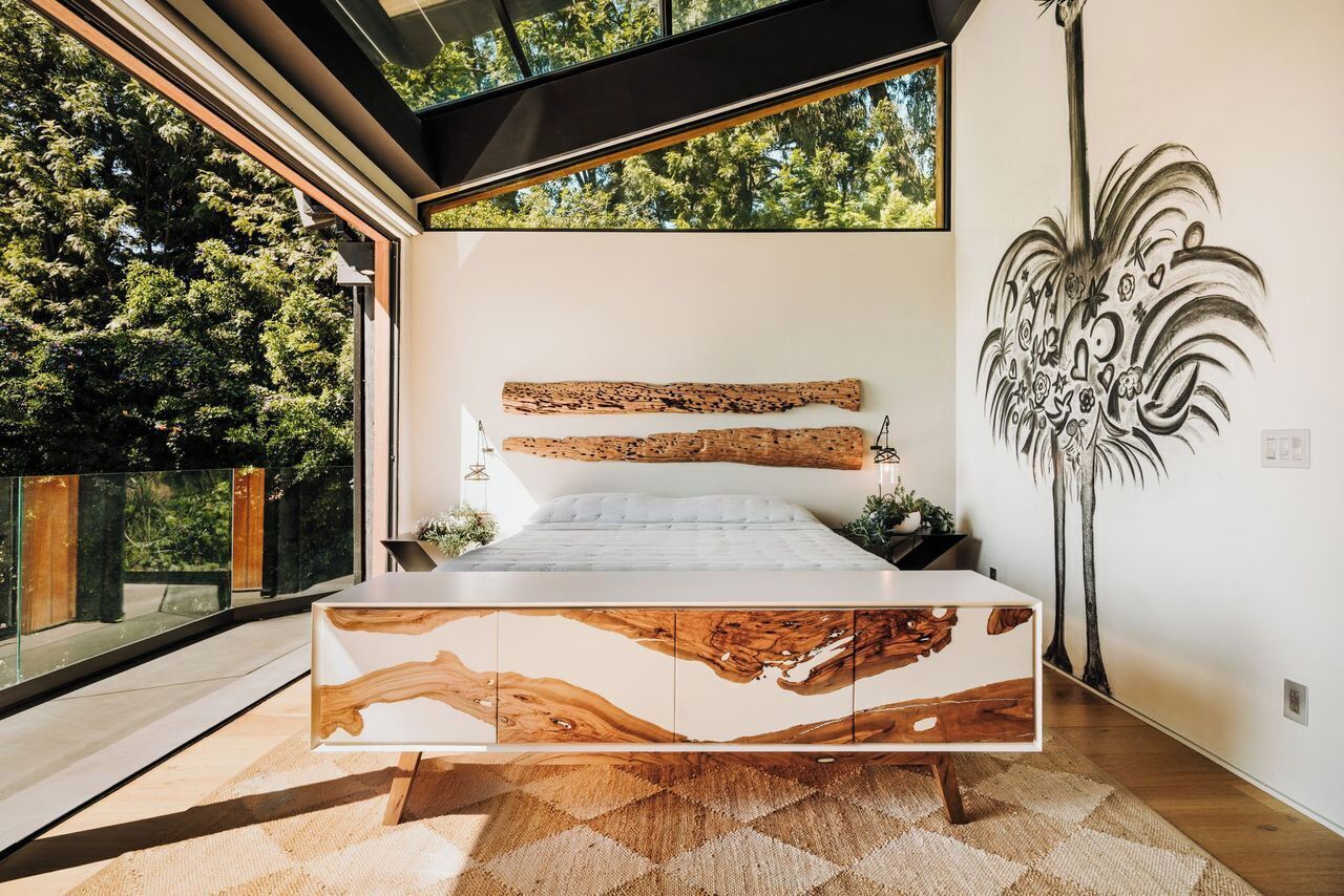 Camera moderna con un letto grande, testiera in legno e vista su alberi attraverso ampie finestre.