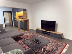 A modern living room with a sofa, a coffee table, a TV, and a kitchen area in the background. - Apartment in the Grafschaft with Shared Terrace, Private Garden, and Wi-Fi (Gelsdorf)