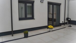 Un edificio con fachada blanca, ventanas de marco negro y una puerta con paneles de vidrio. Hay dos macetas con plantas de flores amarillas.