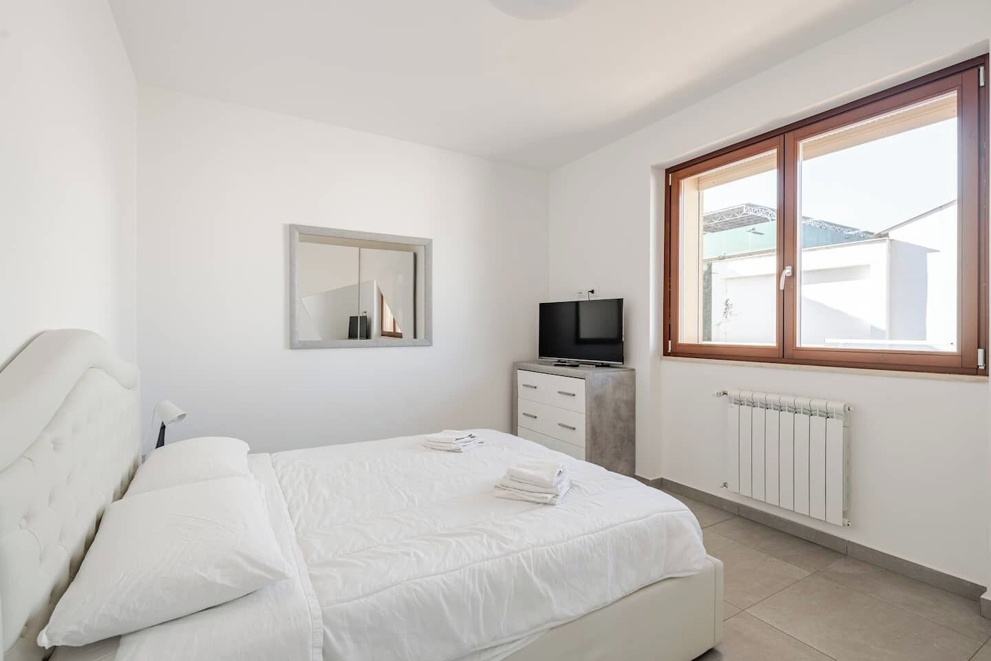 Ein Schlafzimmer mit Bett, Fernseher, Kommode, Spiegel und Fenster mit Blick auf Gebäude.