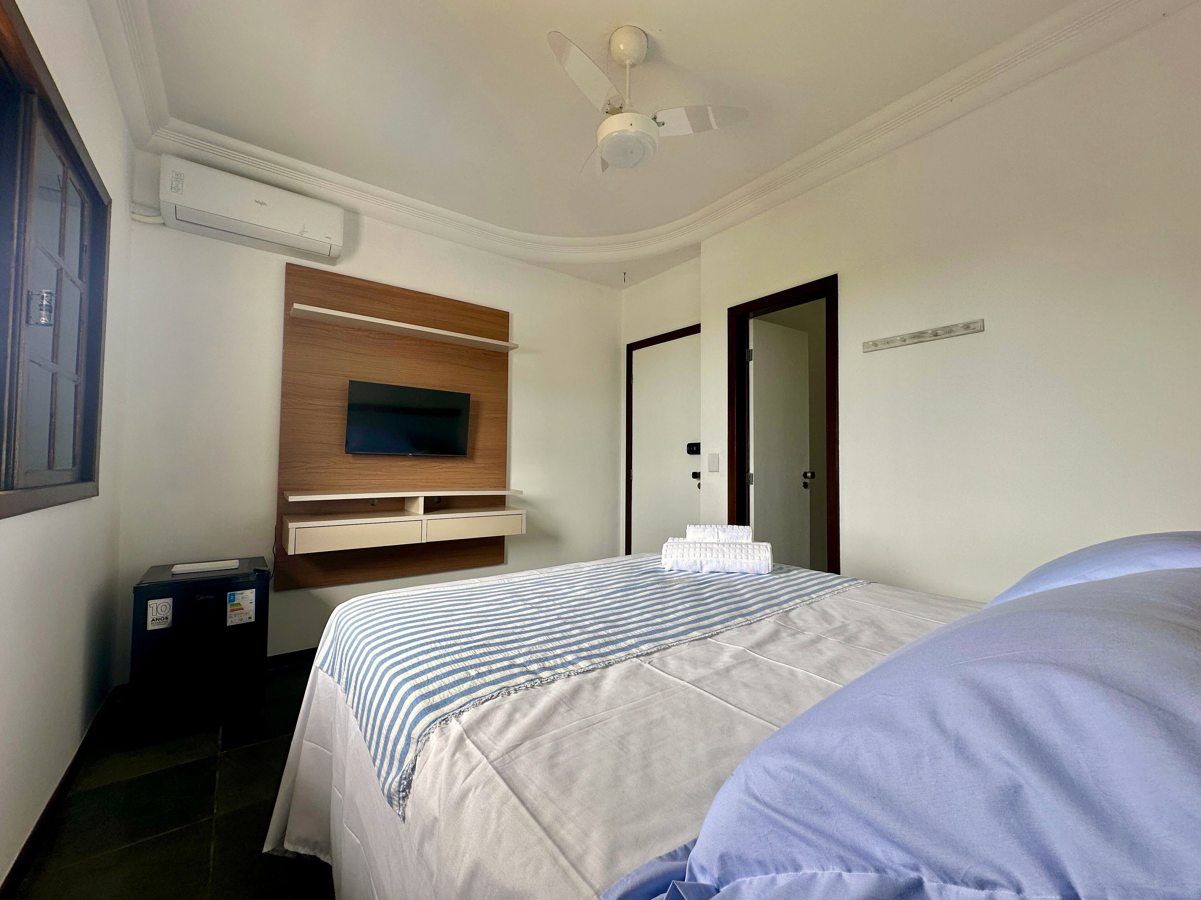 A hotel room with a bed, a TV mounted on a wooden panel, a ceiling fan, and a door.