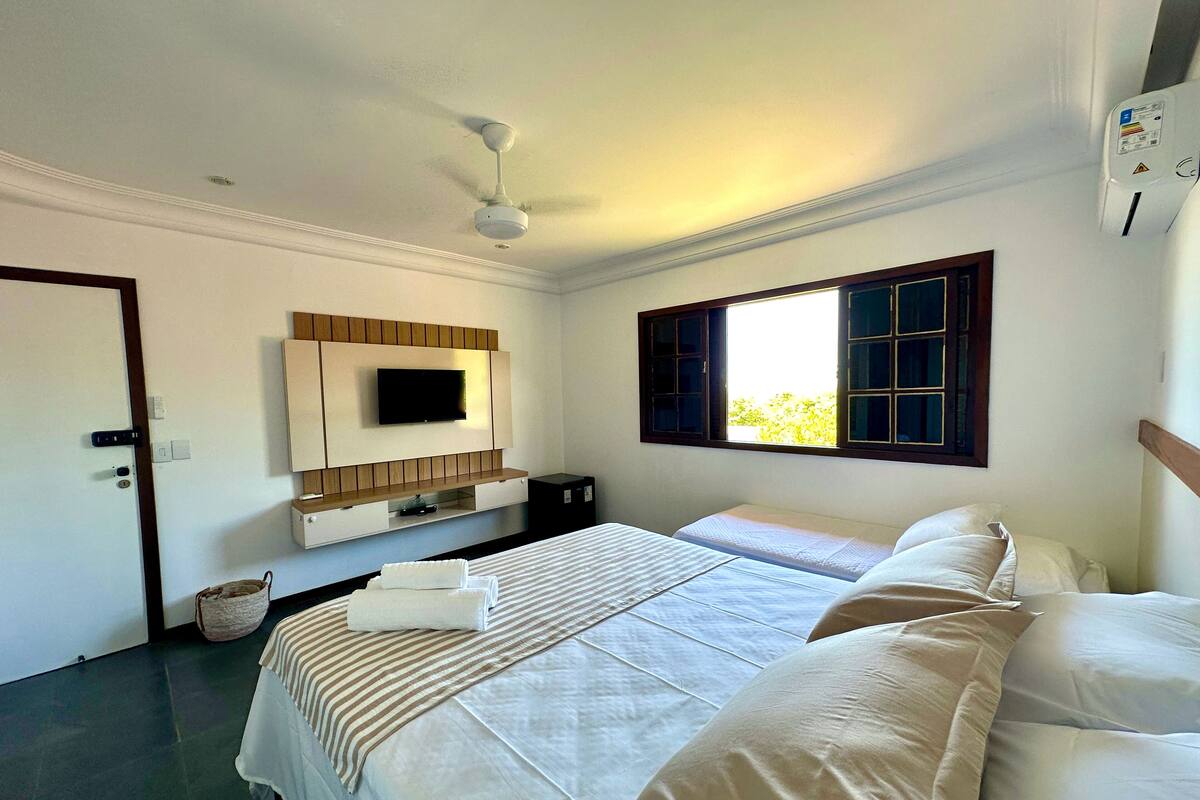 A hotel room with two beds, a television mounted on the wall, a ceiling fan, and a window with a view of greenery outside.