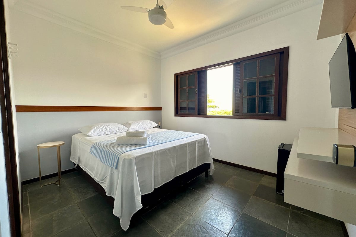 A clean, well-lit hotel room with a bed, a small stool, a window with shutters, and a ceiling fan.