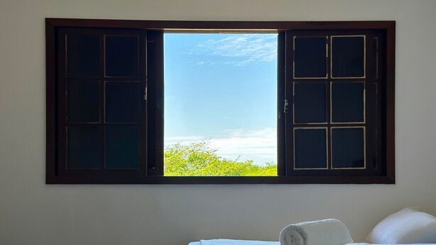 Una stanza con un letto, due asciugamani arrotolati e una finestra vista su vegetazione e cielo.
