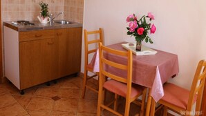 Une cuisine compacte avec des meubles en bois, un évier et un petit plan de travail. On y trouve une table recouverte d’une nappe rose et deux chaises. Un vase rempli de fleurs roses est posé sur la table.