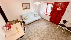 Ein kleiner, gut beleuchteter Raum mit einem Bett, einem Tisch mit Stuhl, einer roten Tür und einem Fenster mit Vorhängen.