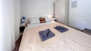 Ein ordentlich gemachtes Bett mit einer beigen Bettdecke, zwei aufgerollten Handtüchern und einem Nachttisch mit einer Lampe.