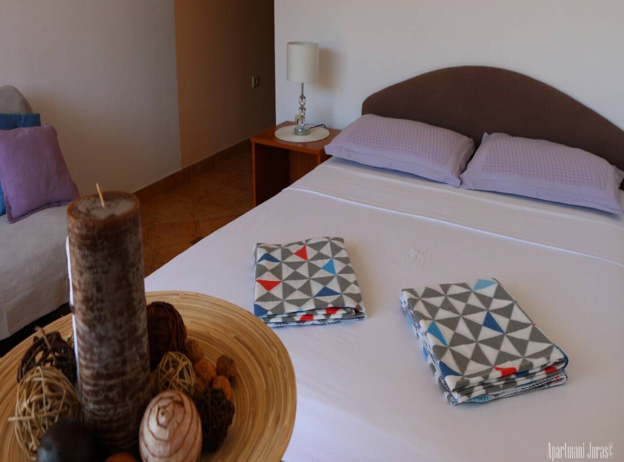 Quarto de hotel com cama, almofadas, mesa de cabeceira com abajur e um objeto decorativo sobre a cama.