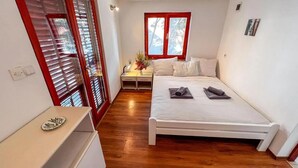 A bedroom with a wooden floor, a bed with pillows and folded towels, a nightstand with a lamp, a window with blinds, and a small table with a decorative item. - Beach front apartment in Gdinj with WiFi (Gdinj)