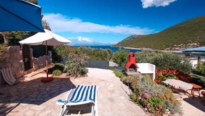 A patio with a stone floor, a striped lounge chair, a red grill, and a view of the sea and hills. - Beach front apartment in Gdinj with WiFi (Gdinj)