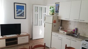 Uma cozinha compacta com armários brancos, geladeira, fogão e pia. Há uma mesa de jantar com cadeiras e uma televisão sobre um suporte.