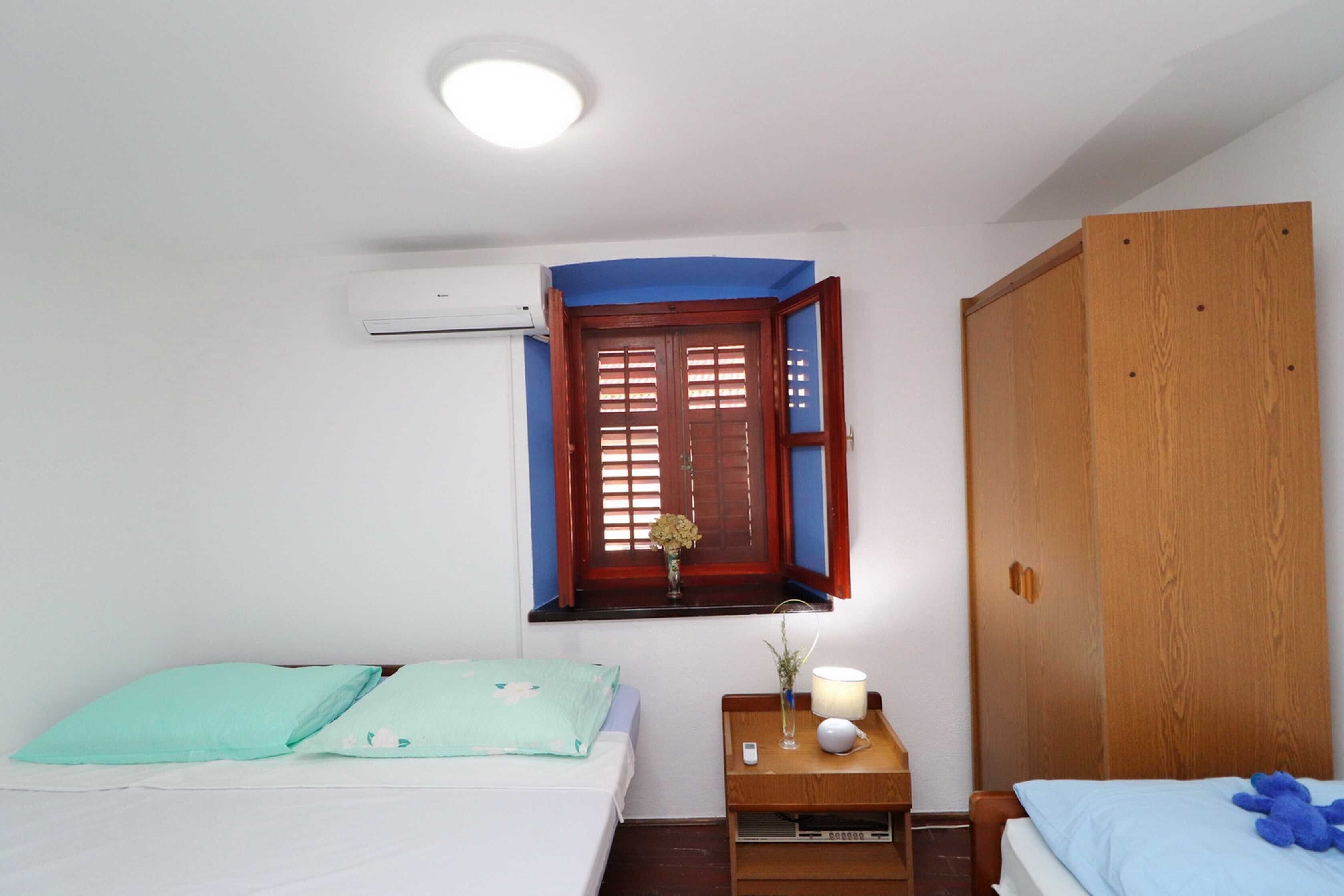 Un dormitorio con cama, mesita de noche, armario de madera, ventana con contraventanas y un equipo de aire acondicionado.