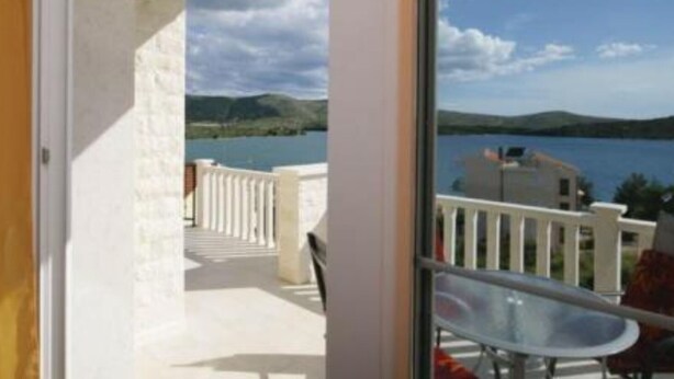 Un balcón con puerta de cristal que da a una terraza con mesa y sillas, con vistas a un cuerpo de agua y colinas.