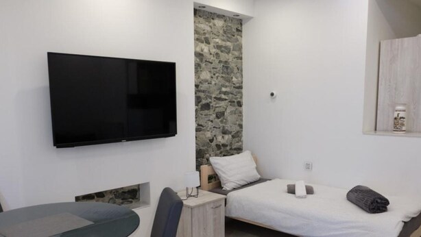 Un dormitorio pequeño y moderno con cama, televisor montado en la pared, una mesita y una silla.