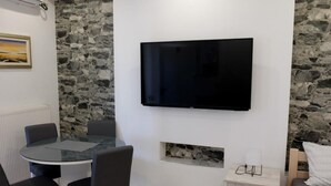 Una habitación moderna con pared de piedra, una mesa redonda con sillas, televisor montado, una estantería pequeña con objetos decorativos y una cama con cabecero.