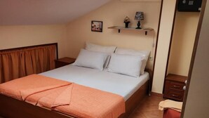 Una camera con un letto singolo, una testiera in legno, una coperta marrone e un materasso azzurro chiaro. È presente un comodino con una lampada e un portafoto. La stanza ha un pavimento piastrellato e una tenda marrone alla finestra.